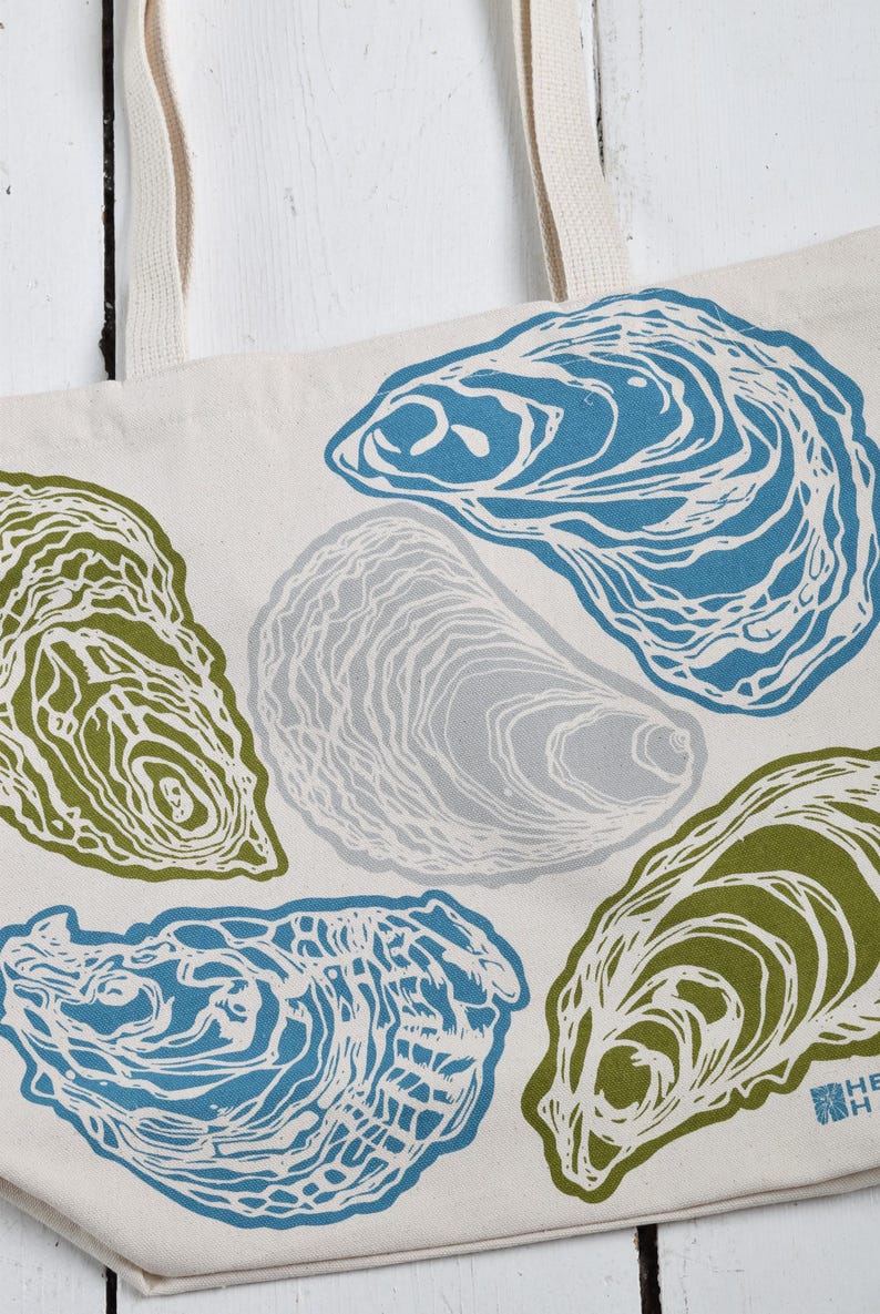 May include: Off-white tote bag featuring a repeating pattern of stylized oyster shells in shades of blue, green, and gray. The bag has cream-colored handles and a simple, clean design, suitable for carrying groceries or personal items.
