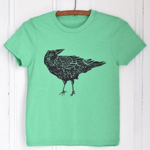 Pode incluir: Uma camiseta verde com um gr&aacute;fico preto de um corvo. O corvo est&aacute; em p&eacute; com as asas ligeiramente abertas. A camiseta est&aacute; pendurada em um cabide de madeira contra um fundo branco.