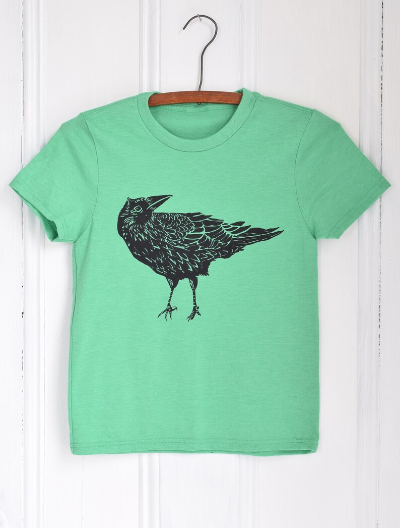 Pode incluir: Uma camiseta verde com um gr&aacute;fico preto de um corvo. O corvo est&aacute; em p&eacute; com as asas ligeiramente abertas. A camiseta est&aacute; pendurada em um cabide de madeira contra um fundo branco.