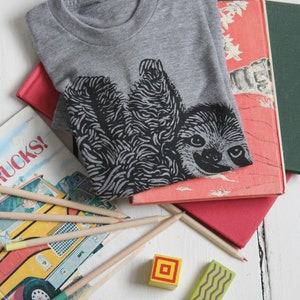 May include: A gray t-shirt with a black and white illustration of a sloth hanging upside down. The shirt is on top of a stack of books.
