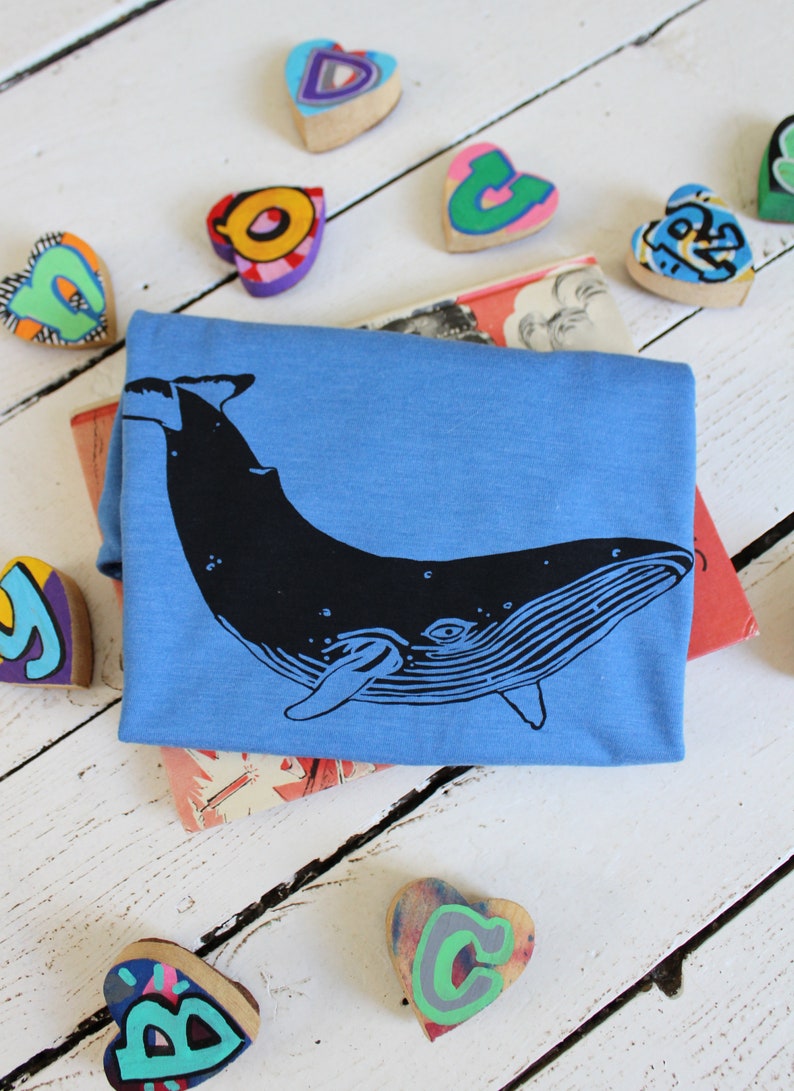 May include: A blue t-shirt with a black whale graphic. The shirt is surrounded by colorful wooden hearts with letters painted on them.