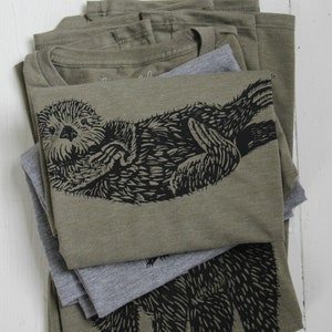 May include: Stack of olive green t-shirts with a black and white graphic print of an otter. The otter is curled up and appears to be sleeping. The t-shirts are folded and stacked on top of each other.