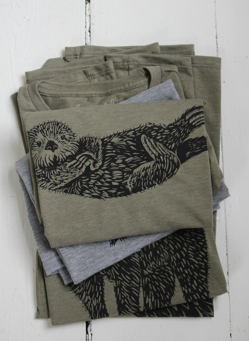 May include: Stack of olive green t-shirts with a black and white graphic print of an otter. The otter is curled up and appears to be sleeping. The t-shirts are folded and stacked on top of each other.