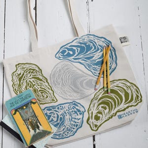 May include: A cream-colored tote bag featuring oyster shell designs in blue, green, and gray. The bag has sturdy cream-colored handles and the words "HEARTH & HARROW" printed on the lower right. A copy of "The Lion, the Witch, and the Wardrobe" and pencils are next to the bag.