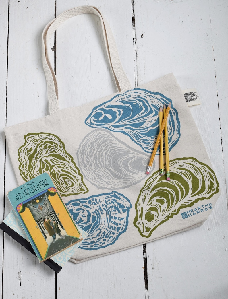 May include: A cream-colored tote bag featuring oyster shell designs in blue, green, and gray. The bag has sturdy cream-colored handles and the words "HEARTH & HARROW" printed on the lower right. A copy of "The Lion, the Witch, and the Wardrobe" and pencils are next to the bag.