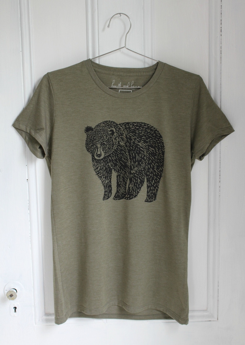 K&ouml;nnte beinhalten: Ein gr&uuml;nes T-Shirt mit einem schwarzen B&auml;ren-Druck. Das Shirt hat einen Rundhalsausschnitt und kurze &Auml;rmel. Der Text "hearth and home" ist auf dem Etikett aufgedruckt.