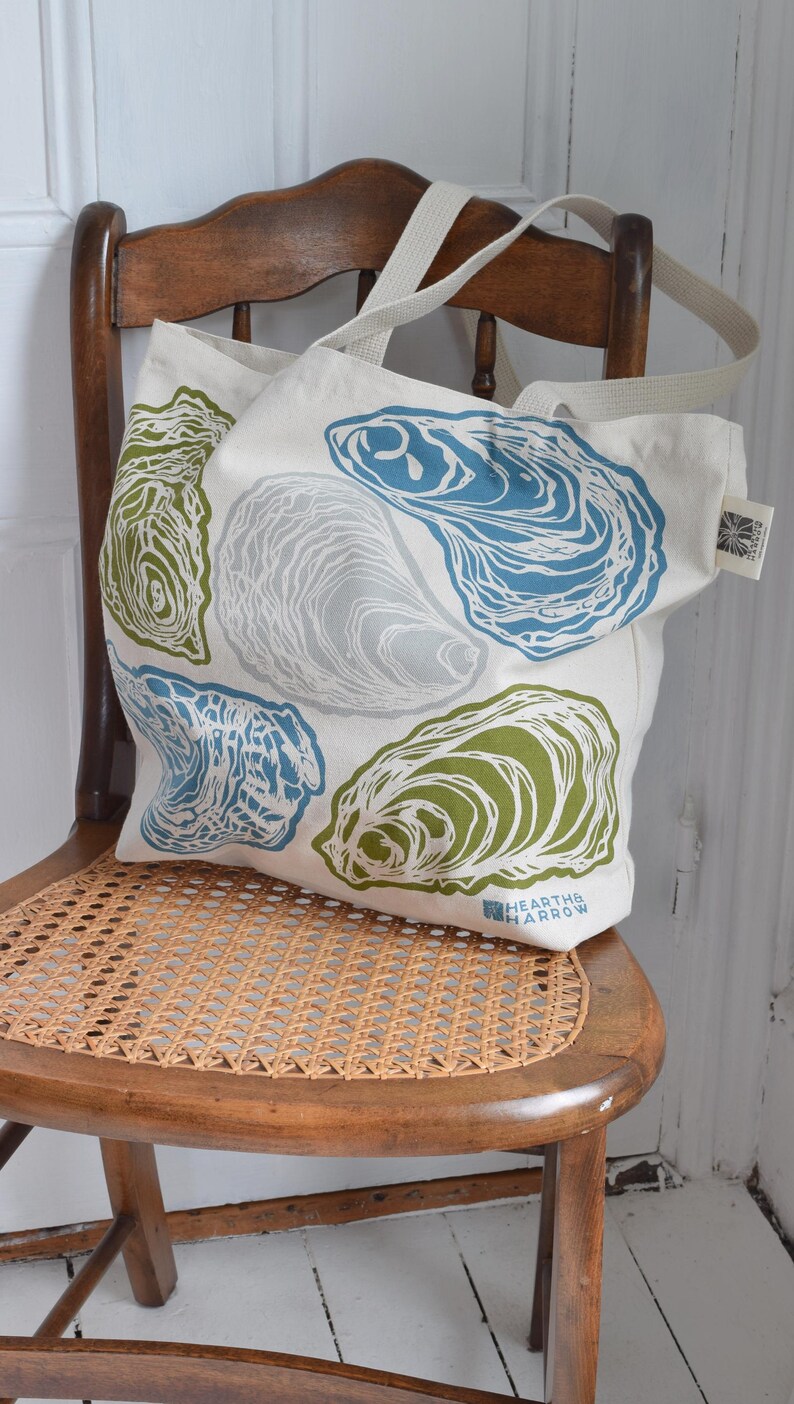 May include: A canvas tote bag with a repeating oyster shell design in blue, green, and gray on a cream background. The bag has cream-colored straps and is resting on a wooden chair.