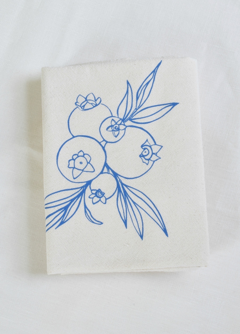 May include: A folded, off-white cloth napkin featuring a blue line drawing of blueberries and leaves. The illustration depicts several blueberries clustered together with delicate stems and foliage, creating a simple, elegant design.