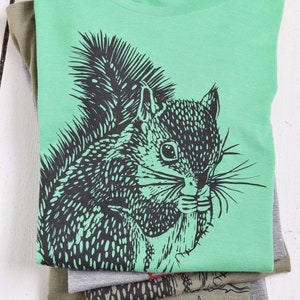 May include: A green t-shirt with a black and white illustration of a squirrel on the front. The squirrel is holding a nut in its mouth.