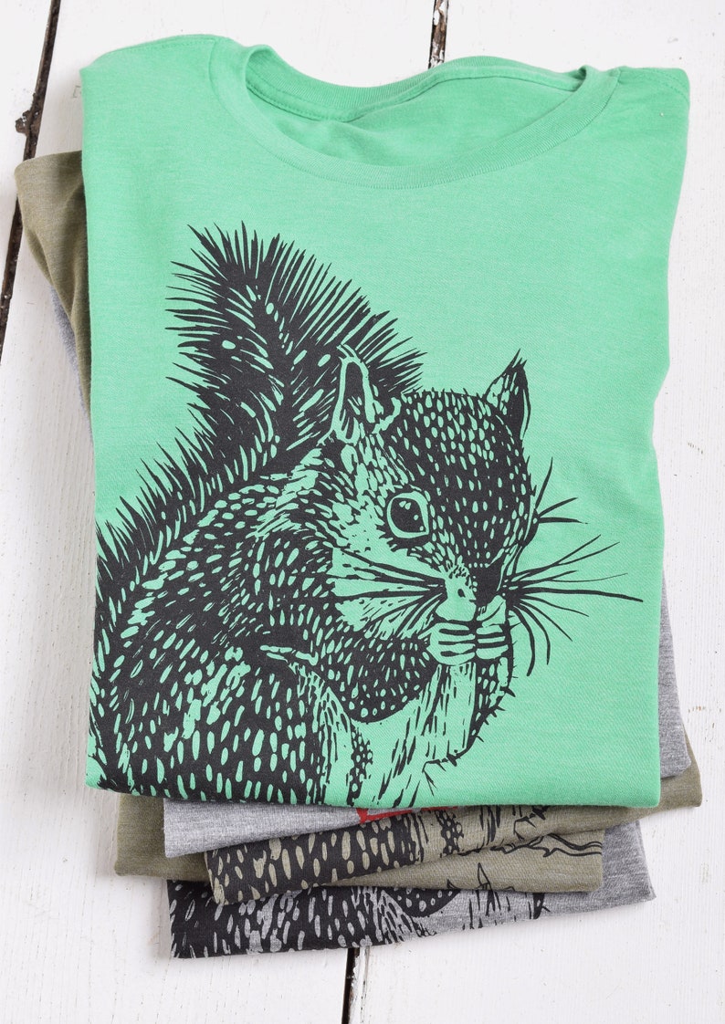 May include: A green t-shirt with a black and white illustration of a squirrel on the front. The squirrel is holding a nut in its mouth.
