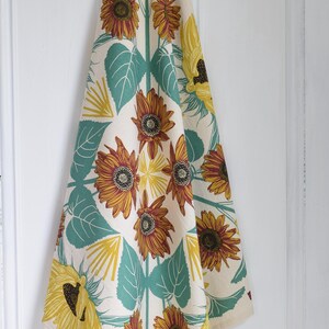 Op de afbeelding: Cr&egrave;mekleurige keukendoek met een zonnebloemmotief. Het ontwerp toont grote gele en bruine zonnebloemen, turquoise bladeren en gele accenten. De handdoek hangt tegen een witte achtergrond.