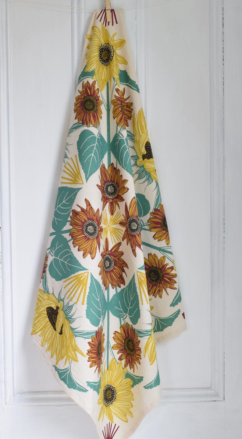 Op de afbeelding: Cr&egrave;mekleurige keukendoek met een zonnebloemmotief. Het ontwerp toont grote gele en bruine zonnebloemen, turquoise bladeren en gele accenten. De handdoek hangt tegen een witte achtergrond.
