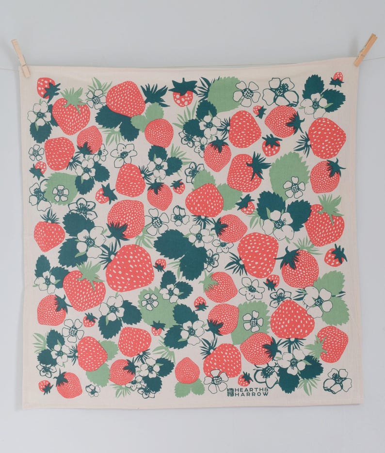 以下が含まれることがあります： 赤いイチゴ、緑の葉、白い花が繰り返し描かれた白いキッチンクロス。右下に「HEARTH & HARROW」の文字が印刷されています。