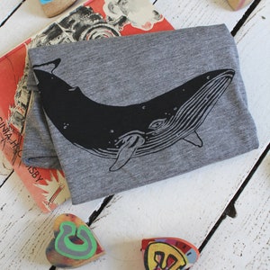 May include: A gray t-shirt with a black whale graphic. The whale is facing right and has a detailed design. The t-shirt is lying on a white wooden surface with colorful heart-shaped objects.
