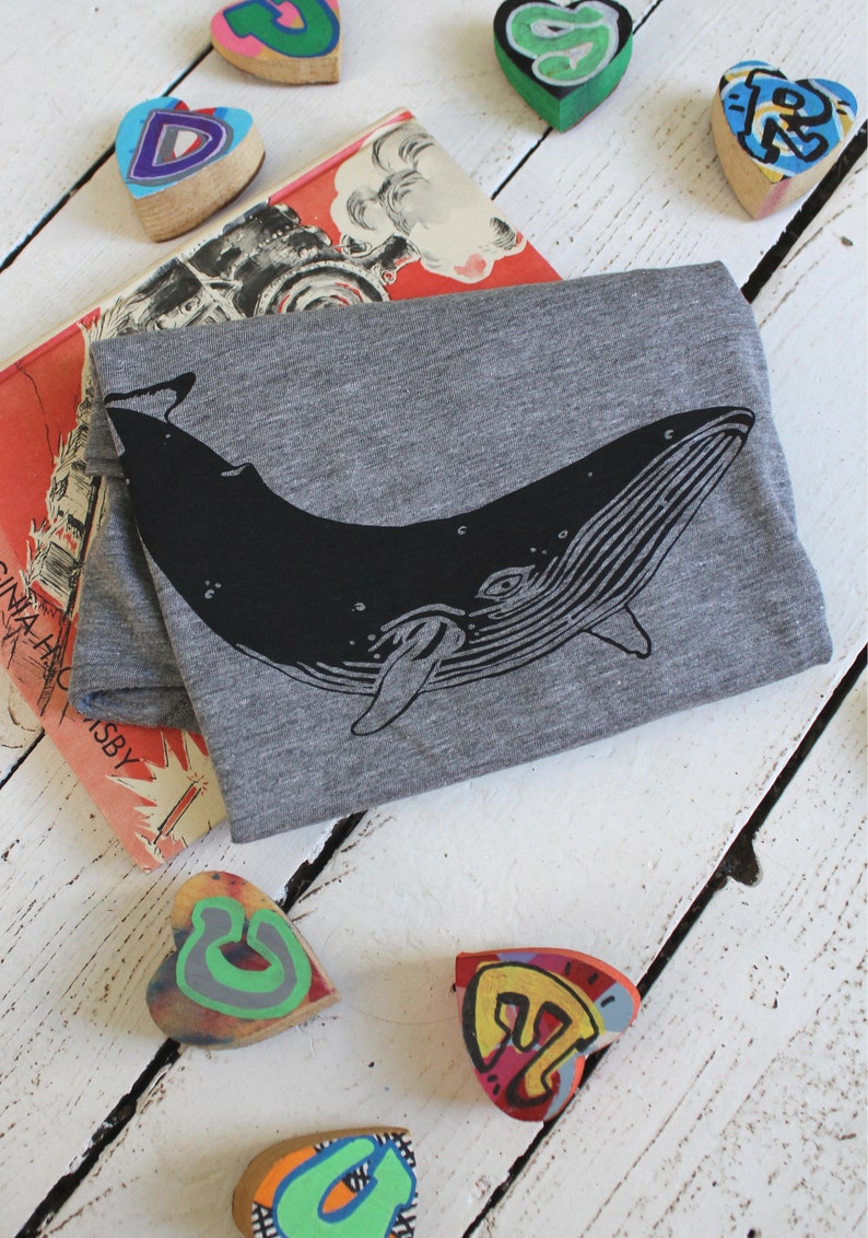 May include: A gray t-shirt with a black whale graphic. The whale is facing right and has a detailed design. The t-shirt is lying on a white wooden surface with colorful heart-shaped objects.