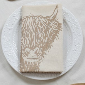 May include: A folded beige linen napkin with a brown Highland cow print rests on a white plate with a floral design. Two wooden spoons are placed on either side of the plate, creating a rustic, farmhouse aesthetic.