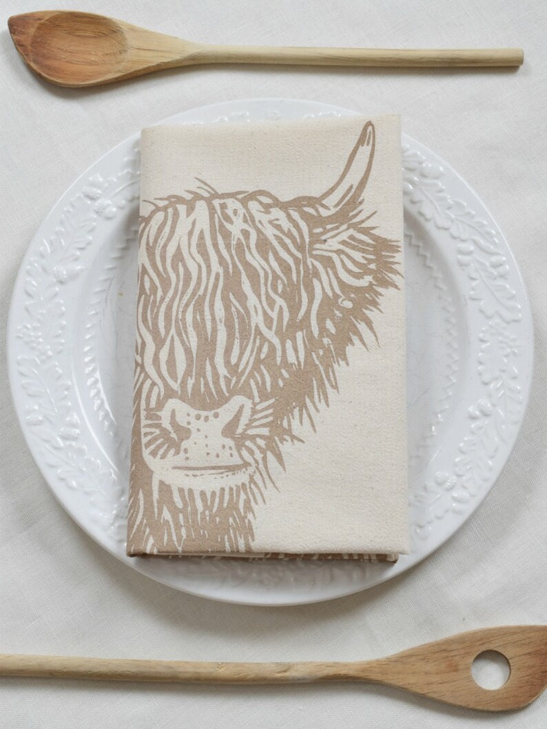 May include: A folded beige linen napkin with a brown Highland cow print rests on a white plate with a floral design. Two wooden spoons are placed on either side of the plate, creating a rustic, farmhouse aesthetic.