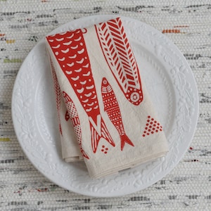 May include: A white ceramic plate with a raised floral design around the edge. A folded red and white linen napkin with a red fish design is on the plate.