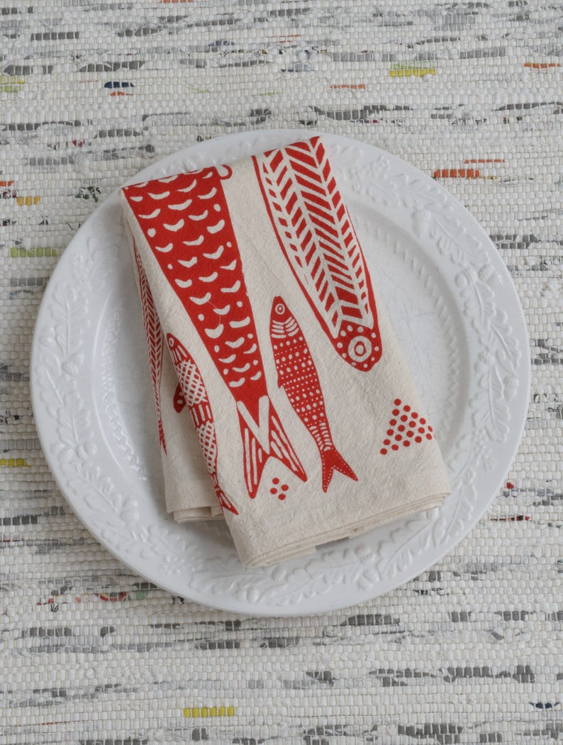 May include: A white ceramic plate with a raised floral design around the edge. A folded red and white linen napkin with a red fish design is on the plate.