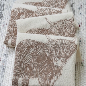 May include: Four cream-colored tea towels with a brown Highland cow design. The towels are stacked on top of each other.