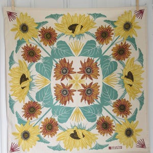 Op de afbeelding: Cr&egrave;mekleurig vierkant textiel met een herhalend zonnebloemontwerp. De zonnebloemen zijn geel en bruin, met turquoise bladeren en accenten. Het textiel heeft een geometrisch patroon en de woorden "HEARTH & HARROW" staan rechtsonder gedrukt.