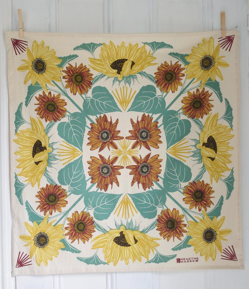 Op de afbeelding: Cr&egrave;mekleurig vierkant textiel met een herhalend zonnebloemontwerp. De zonnebloemen zijn geel en bruin, met turquoise bladeren en accenten. Het textiel heeft een geometrisch patroon en de woorden "HEARTH & HARROW" staan rechtsonder gedrukt.