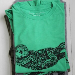 K&ouml;nnte beinhalten: Ein Stapel gefalteter T-Shirts in Gr&uuml;n- und Graut&ouml;nen. Das oberste Shirt ist leuchtend gr&uuml;n und zeigt eine schwarze Grafik einer Seeotter. Der Text "heart and narrow" ist auf dem Etikett zu sehen. Die Shirts sind auf einer wei&szlig;en Holzoberfl&auml;che gestapelt.
