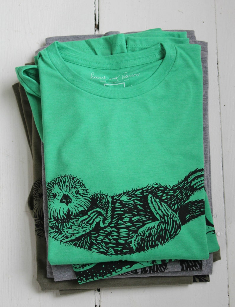 K&ouml;nnte beinhalten: Ein Stapel gefalteter T-Shirts in Gr&uuml;n- und Graut&ouml;nen. Das oberste Shirt ist leuchtend gr&uuml;n und zeigt eine schwarze Grafik einer Seeotter. Der Text "heart and narrow" ist auf dem Etikett zu sehen. Die Shirts sind auf einer wei&szlig;en Holzoberfl&auml;che gestapelt.