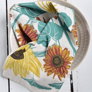 Op de afbeelding: Een gevouwen beige keukendoek met een zonnebloemmotief, met gele en bordeauxrode zonnebloemen met turquoise bladeren. De handdoek ligt op een beige bord met een geribbeld ontwerp, op een wit houten oppervlak.
