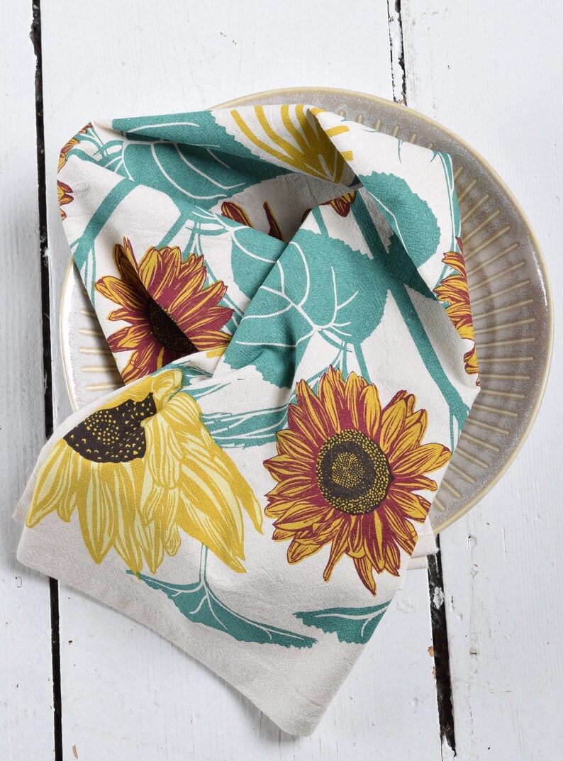 Op de afbeelding: Een gevouwen beige keukendoek met een zonnebloemmotief, met gele en bordeauxrode zonnebloemen met turquoise bladeren. De handdoek ligt op een beige bord met een geribbeld ontwerp, op een wit houten oppervlak.