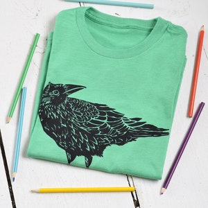 Pode incluir: Uma camiseta verde-claro dobrada com um desenho preto de um corvo. V&aacute;rios l&aacute;pis de cor, incluindo azul, verde, amarelo e vermelho, est&atilde;o espalhados ao redor da camiseta em uma superf&iacute;cie de madeira branca. O desenho do corvo &eacute; detalhado.