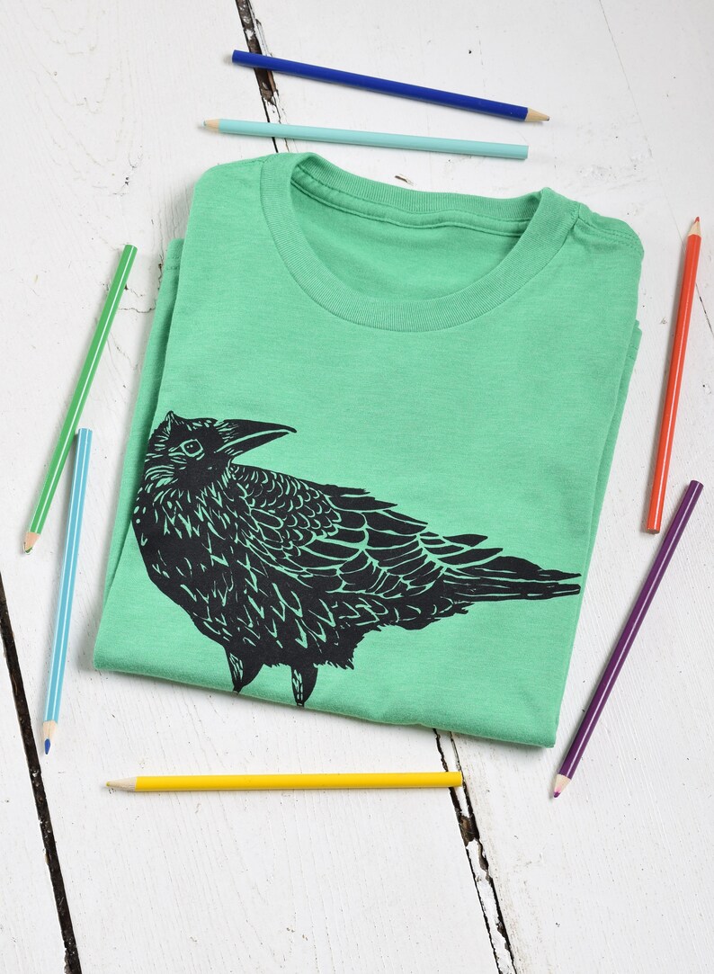 Pode incluir: Uma camiseta verde-claro dobrada com um desenho preto de um corvo. V&aacute;rios l&aacute;pis de cor, incluindo azul, verde, amarelo e vermelho, est&atilde;o espalhados ao redor da camiseta em uma superf&iacute;cie de madeira branca. O desenho do corvo &eacute; detalhado.