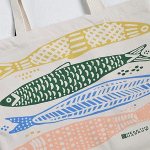 May include: Cream-colored tote bag featuring a colorful fish design. The fish are yellow, green, blue, and peach, with various patterns. The bag has cream-colored handles and the Hearth & Harrow logo.