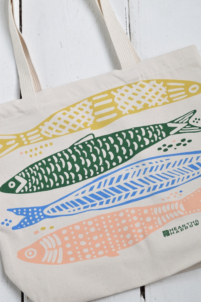 May include: Cream-colored tote bag featuring a colorful fish design. The fish are yellow, green, blue, and peach, with various patterns. The bag has cream-colored handles and the Hearth & Harrow logo.