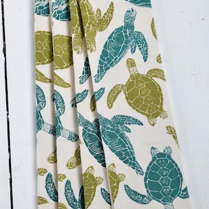 May include: Four folded cream-colored kitchen towels with a repeating pattern of sea turtles in teal and olive green. The turtles are stylized with detailed shell patterns, creating a coastal theme.