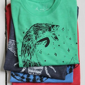 Puede incluir: Pila de cuatro camisetas con diferentes dise&ntilde;os. La camiseta superior es verde con una ilustraci&oacute;n de zorro negro y el texto "Earth and Hand" en la etiqueta. La segunda camiseta es azul oscuro con una ilustraci&oacute;n de insecto blanco. La tercera camiseta es azul con una ilustraci&oacute;n de insecto negro. La camiseta inferior es roja.