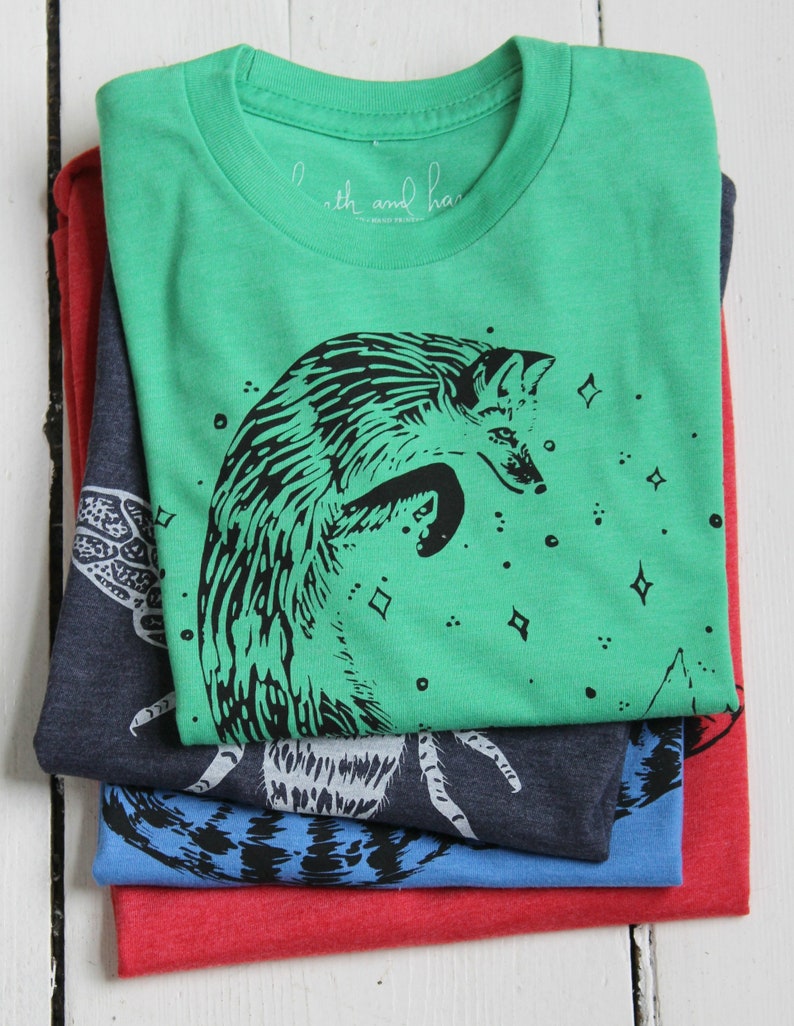 Puede incluir: Pila de cuatro camisetas con diferentes dise&ntilde;os. La camiseta superior es verde con una ilustraci&oacute;n de zorro negro y el texto "Earth and Hand" en la etiqueta. La segunda camiseta es azul oscuro con una ilustraci&oacute;n de insecto blanco. La tercera camiseta es azul con una ilustraci&oacute;n de insecto negro. La camiseta inferior es roja.