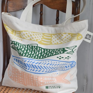 May include: Cream-colored tote bag with four colorful fish designs in yellow, green, blue, and peach. The bag has cream-colored handles and the text "HEARTH & HARROW" printed on the bottom.