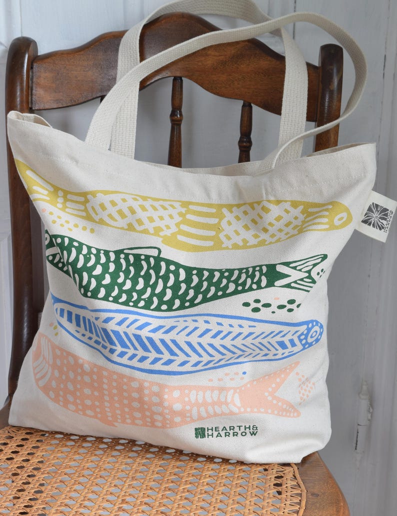 May include: Cream-colored tote bag with four colorful fish designs in yellow, green, blue, and peach. The bag has cream-colored handles and the text "HEARTH & HARROW" printed on the bottom.