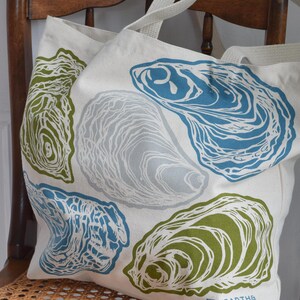 May include: A cream-colored tote bag featuring a repeating pattern of oyster shell illustrations in shades of blue, green, and gray. The bag has sturdy cream-colored straps and the Hearth & Harrow logo on the bottom right corner. The bag is sitting on a wooden chair.