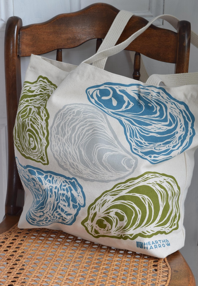 May include: A cream-colored tote bag featuring a repeating pattern of oyster shell illustrations in shades of blue, green, and gray. The bag has sturdy cream-colored straps and the Hearth & Harrow logo on the bottom right corner. The bag is sitting on a wooden chair.