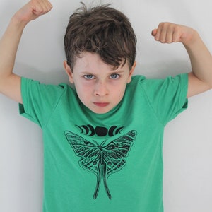 May include: A young boy wearing a green t-shirt with a black and white butterfly graphic and a crescent moon design. The boy is flexing his biceps.