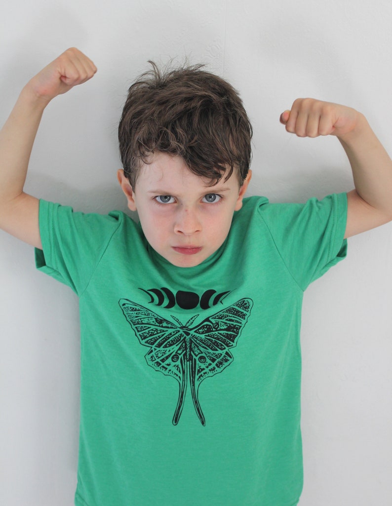 May include: A young boy wearing a green t-shirt with a black and white butterfly graphic and a crescent moon design. The boy is flexing his biceps.