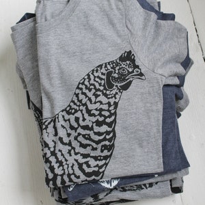 May include: A stack of gray and navy blue t-shirts with a black and white graphic print of a chicken on the top shirt.