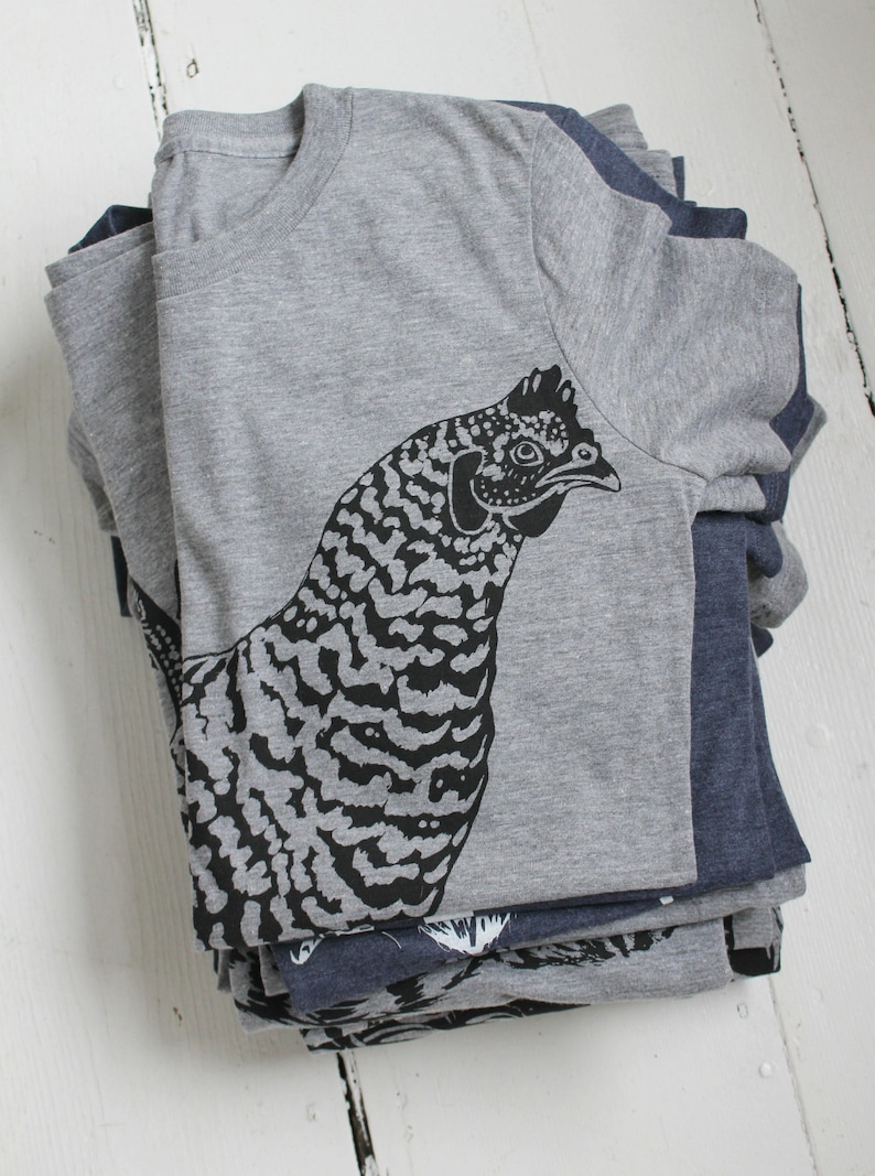 May include: A stack of gray and navy blue t-shirts with a black and white graphic print of a chicken on the top shirt.