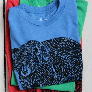 May include: A stack of three t-shirts, one red, one green, and one blue. The blue t-shirt has a black and white illustration of a bear on the front. The text "earth and" is visible on the blue t-shirt.