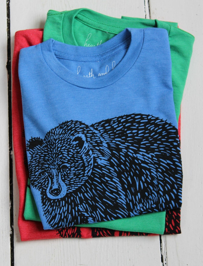 May include: A stack of three t-shirts, one red, one green, and one blue. The blue t-shirt has a black and white illustration of a bear on the front. The text "earth and" is visible on the blue t-shirt.