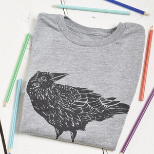 Pode incluir: Uma camiseta cinza com um gr&aacute;fico preto de um corvo. A camiseta est&aacute; dobrada e rodeada por l&aacute;pis de cor. O desenho do corvo &eacute; detalhado, com penas vis&iacute;veis e um bico afiado. Os l&aacute;pis s&atilde;o de v&aacute;rias cores, incluindo azul, verde e amarelo.