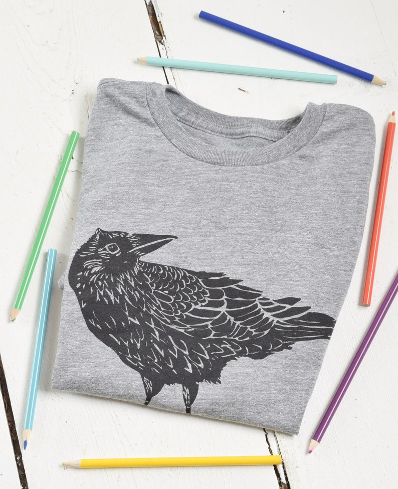 Pode incluir: Uma camiseta cinza com um gr&aacute;fico preto de um corvo. A camiseta est&aacute; dobrada e rodeada por l&aacute;pis de cor. O desenho do corvo &eacute; detalhado, com penas vis&iacute;veis e um bico afiado. Os l&aacute;pis s&atilde;o de v&aacute;rias cores, incluindo azul, verde e amarelo.