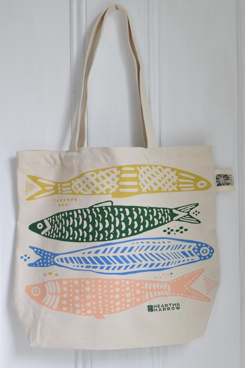 May include: A beige tote bag featuring four colorful fish illustrations in yellow, green, blue, and peach. The fish designs are stylized with various patterns. The bag has long beige handles and the text "HEARTH & HARROW" is printed on the bottom.